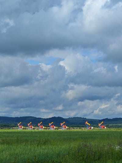 In a rural landscape, a line of seven red and orange oil pumpjacks stretches horizontally across the mid-ground of the image. They are positioned in front of a backdrop of rolling green hills and a dark, cloudy sky. In the foreground, a field of tall green grass and some flowering plants occupies the lower portion of the frame. The image is a landscape photograph taken from a distance, capturing the industrial equipment against a natural setting. The location is specified as Kipchak, Russia.
