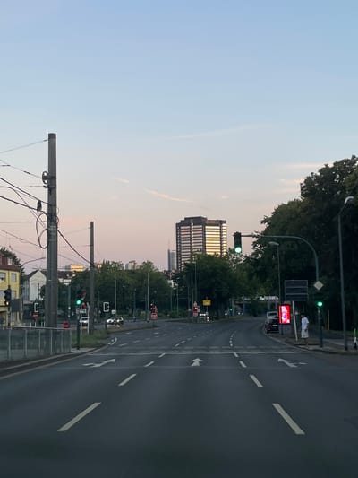 The image shows a view of a city street in Essen, Germany, at dusk. The sky is a gradient of pale blue and pink. In the foreground, a multi-lane asphalt road is visible with white lane markings and arrows indicating traffic flow. Several cars are on the road, some moving and some stopped at traffic lights. There are traffic lights, street signs, and utility poles lining the street.
To the left, a building with yellow facade is visible, and a tram or light rail track runs along the side of the road. Further down the street, a tall, modern office building with a distinctive glass facade dominates the skyline, reflecting the warm colors of the sunset. Lush green trees are abundant on both sides of the road, creating a contrast with the urban environment.
On the right side of the road, a man in a white t-shirt and shorts stands on the sidewalk next to a large digital advertising screen. A few parked cars are also visible. The overall scene depicts a typical urban streetscape as day transitions into night, with the city lights beginning to appear on some of the buildings.