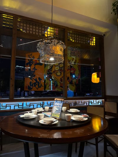 The image captures an indoor setting in Zhangjiajie, China, likely a restaurant. A round, dark wood table is set with white ceramic tea cups and saucers, and chopsticks, suggesting a tea service. A sign in Chinese is positioned at the table. The backdrop features a large window showcasing decorative Chinese artwork and city lights. A modern, metallic chandelier hangs above the table. The overall lighting is a mix of natural light from the window and artificial illumination from the chandelier and a distant yellow lantern visible outside. The environment appears upscale with intricate woodwork and attention to detail.