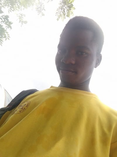 A medium-shot, low-angle, underexposed, outdoor image shows a person wearing a yellow shirt with a patch on the left breast and a black backpack on their left shoulder. The person has dark skin, short hair, and is looking at the camera with a slight smile. The background is a bright, overcast sky with some green leaves visible at the top of the frame. The location is Eleweran, Nigeria.