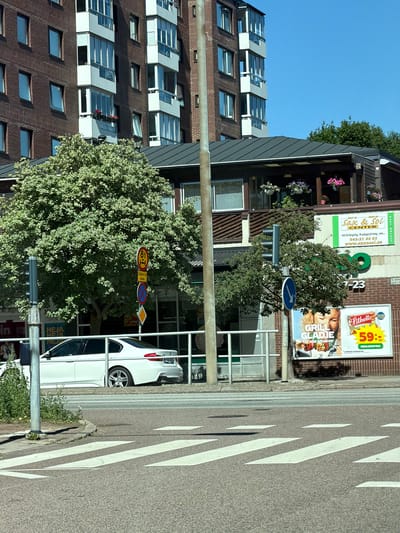 This is a medium shot, taken from a low angle, of a street scene in Helsingborg, Sweden. The image is oriented vertically, with the sky at the top and the street at the bottom. The scene features a multi-story building on the left, with several floors of windows and balconies. A large, leafy green tree partially obscures the building and the street. In the foreground, a white car is parked on the right side of the street, next to a pedestrian crossing marked with white stripes.
Several signs are visible on the building. One sign for "Sax & Sol CENTER" advertises hair extensions and bridal styling, with a phone number and website. Another sign displays "Lithells" and a promotion for "GRILLA GLÄDJE för alla smaker" (Grilling Joy for all tastes) with a price of "59:-/st". There are also traffic signs, including a speed limit sign (8t) and other regulatory signs. The building appears to be a commercial establishment, possibly a salon or shop, with apartments above. The weather appears to be sunny, with a clear blue sky, suggesting it is daytime. The scene depicts a typical urban street environment with a mix of commercial and residential architecture, and common street elements like cars and traffic signs.