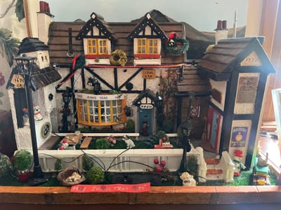 The image depicts a handcrafted, miniature replica of a Tudor-style building, likely a commemorative piece relating to "Jean's Teas." It features a main building with two dormer windows, decorated with wreaths and holiday embellishments. The architecture is characteristic of Tudor style, with dark timber framing against white walls. In front of the main building, a white picket fence encloses a miniature garden scene with toy animals, faux greenery, and a small table. To the left and right of the main structure are smaller, attached buildings with similar architectural elements and additional detailing such as miniature street lamps and advertisements. A sign indicates "1935 Jean's Teas 1995," suggesting a historical connection. The model sits on a base that mimics a sidewalk. A backdrop of rolling hills implies a setting outside of a city. The city location of Golspie, United Kingdom, is not visible in this image.