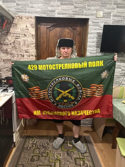 A man stands indoors, holding a flag. The flag is divided vertically into green and red sections, with the text "429 мотострелковый полк" on the green side and "ИМ. Кубанского казачества" on the red side, both written in yellow. The flag also features the ribbon of Saint George and a logo that contains crossed rifles. The man is wearing a camouflage-patterned jacket and a black hat. He appears to be in a room, with a wooden door behind him and patterned wallpaper. The location is Vesyoloye, Russia.