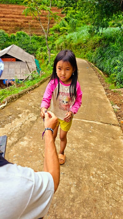 The image captures a ground-level perspective of a young Asian girl interacting with an adult male's hand on a worn path in Pung Luong, Vietnam.

The girl, appearing to be between 5-8 years old, stands slightly off-center to the right, looking directly forward with a neutral, serious expression. She has long, straight dark hair and wears small, light-colored dangle earrings. Her attire consists of a long-sleeved shirt with pink sleeves and a light brown torso featuring a partial graphic including the text "TENM" and a cartoon character (possibly a unicorn or pegasus). She wears yellow shorts and light brown, open-toe sandals. Her right hand is extended, gently touching a purple string or bracelet that is held by the outstretched hand and forearm of an adult male, visible in the lower left foreground.

The adult male's forearm is tan and lightly haired, wearing a white short-sleeved shirt. A blue and white patterned bracelet is visible on his wrist, in addition to the purple string being held. A portion of a dark-colored mobile phone or device is visible on the ground in the immediate lower-left corner.

The setting is a rough, light brown concrete or packed earth path exhibiting visible cracks and uneven texture. The path recedes into the distance, curving slightly to the right. To the left of the path, a small, rustic dwelling with a gray corrugated roof and light-colored walls is partially visible, with a blue tarp or structure behind it. The background features lush green tropical vegetation on rolling hillsides. On the left hillside, distinct reddish-brown agricultural terraces are carved into the landscape. Dense green foliage lines both sides of the path as it extends into the midground.