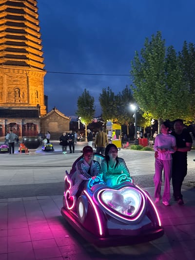 A nighttime scene in Jinzhou, China shows a brightly lit neon ride being enjoyed by a child and an adult. The ride, with pink and blue neon lights, is on a paved area with other people strolling nearby. In the background, lit buildings include a tall, tiered traditional Chinese building. Above, the sky transitions from blue to a darker hue. A couple, a man in black and a woman in pink, are visible in the upper part of the image.