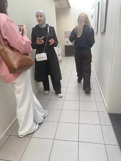 The image depicts an indoor hallway with tiled flooring. In the foreground, a person wearing a pink and white striped shirt and white wide-leg pants stands with their back partially to the camera, holding a brown tote bag. To their right, a woman wearing a black trench coat and a light grey hijab is talking and holding a mobile phone. She has a small white quilted handbag with a gold chain strap. Further down the hallway, a third person with long blonde hair, wearing a dark long-sleeved shirt and brown pants, walks away from the camera. The environment suggests a commercial or public building.