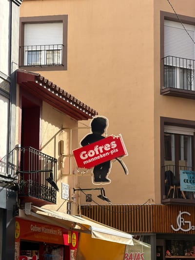 This is an outdoor, slightly angled, eye-level medium shot of a building facade in Benidorm, Spain. The building has two stories visible. The upper story features two windows with white blinds and decorative metal grilles. The wall is a light beige color.

A bright red, illuminated sign is prominently displayed, advertising "Gofres Manneken Pis". The sign has a stylized image of a person and the text is in white. Below this sign, a part of a business is visible with an awning and a red facade. Text on this section also says "Gofres Manneken Pis" and "Productos Naturales y Artesanales".

Further up the facade, above the "Gofres Manneken Pis" sign, there are more signs. One is a light blue sign with white text that reads "APARTAME LA GLOF" and includes what appears to be a reservation phone number and website. Another sign is a white rectangular sign with text in Spanish and a coat of arms, possibly indicating a historical building or a local designation. To the right of these signs, a wooden slatted architectural element is visible, with a white logo that appears to be "GRJ".

On the lower level, a section of a street or alleyway is visible, with a black metal railing and what appears to be more signage or a shop front. The lighting suggests it is daytime, with strong sunlight casting shadows. The overall impression is of a commercial street in a Spanish town.