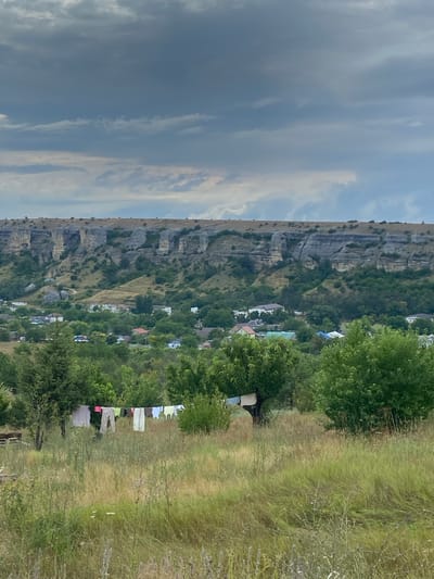 In Krasnomakskoye village, Ukraine, a rural landscape unfolds under a cloudy sky. In the foreground, tall grasses and scattered wildflowers create a natural meadow. A clothesline stretches across the middle ground, adorned with a variety of laundry items, including shirts, pants, and undergarments, fluttering in the breeze. To the left of the clothesline, a rustic bench is partially visible. Beyond this, a cluster of trees provides shade. In the mid-ground, a village is nestled amongst lush greenery, with houses featuring red and blue roofs interspersed with white structures. The background is dominated by a dramatic, weathered cliff face with distinct rock formations, characteristic of the Crimean Mountains. The overall scene captures a tranquil, everyday moment in a rural Ukrainian setting.