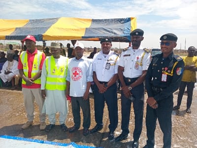 The image shows a group of six men standing under a yellow and blue striped tent. The men are wearing various uniforms, including red and white vests with a red cross symbol, green and yellow vests, white shirts with blue and red stripes, and dark blue military uniforms with yellow and black stripes. The man in the middle is wearing a white shirt with a red cross symbol and a white vest. The man on the far left is wearing a red shirt with a white vest. The men are standing on a paved surface that is wet and appears to be recently cleaned. There are other people in the background, blurred, who appear to be gathered around the tent. The scene is likely taking place in a public space, possibly a plaza or a park. It is taking place in Katabu, Nigeria, at coordinates (latitude: 10.71, longitude: 7.514).