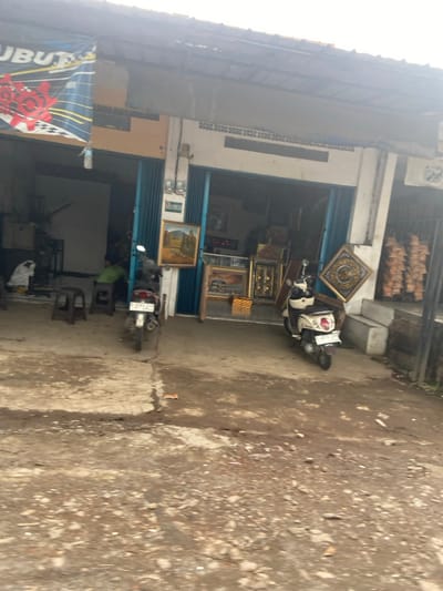 The image shows the exterior of a storefront in Baleendah, Indonesia, located at coordinates -7.021 latitude and 107.656 longitude. Two motorcycles are parked in front of the store, one black and one white. The black motorcycle is partially obscured by a blue door. The white motorcycle is parked in front of a white wall. There is a sign with the word "UBUT" on it. The interior of the storefront is visible, and there are several framed pictures on the walls. Two people are visible in the interior: one is sitting on a chair, and the other is standing. The ground is covered in gravel and dirt, and there are a few empty plastic chairs in front of the store. The building is made of concrete and has a metal roof. There are several hanging objects that appear to be dried fruits. The entrance to the building is open, revealing a large open space. There is a metal door on the right side of the image, and an object is hanging from it. The street is empty, except for the two motorcycles. The sky is overcast and gray. The image is taken from a slightly elevated angle. The image is well lit and in focus.