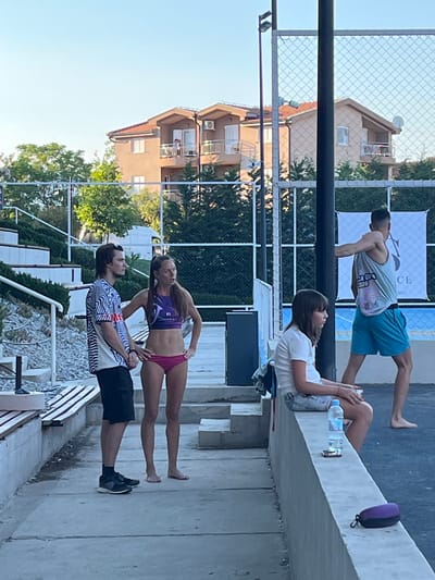 The image captures a group of people at an outdoor sports facility, likely a beach volleyball court, in Gošići, Montenegro. The setting appears to be a sunny day, with clear blue skies. In the foreground, a man and a woman are standing side-by-side, appearing to be in conversation. The man is wearing a black and white patterned shirt and black shorts with athletic shoes. The woman is wearing a purple sports bra and pink bikini bottoms. To the right, a young girl is seated on a concrete ledge, looking to her left. She is wearing a white t-shirt and patterned shorts. Beside her on the ledge are a bottle of water and a mobile phone. Further to the right, a man in a light blue tank top and blue shorts is leaning against a pole, facing away from the camera. In the background, there are multi-story buildings with red-tiled roofs, surrounded by lush green trees. A net and poles suggest a sports court, and a white banner with text is visible behind the man leaning on the pole. The ground is paved with concrete.