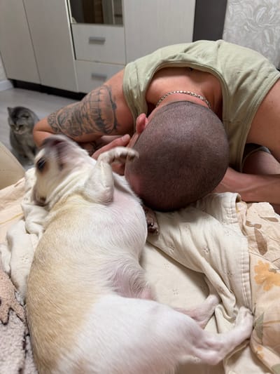 In Semiluki, Russia, a man with tattoos on his left arm is resting on a bed next to a small dog. The man is wearing a light-colored tank top and a silver chain necklace. His head is bowed, and he is nuzzling the dog, which appears to be a small breed with light-colored fur. The dog is lying on its back with its paws in the air, suggesting it is either playfully interacting with the man or is in motion. In the background, a gray cat is visible, sitting on the floor and looking towards the pair. The environment appears to be a bedroom, with a wardrobe and a wall in the background. The bedding is light-colored with a subtle pattern. The lighting suggests it is daytime.