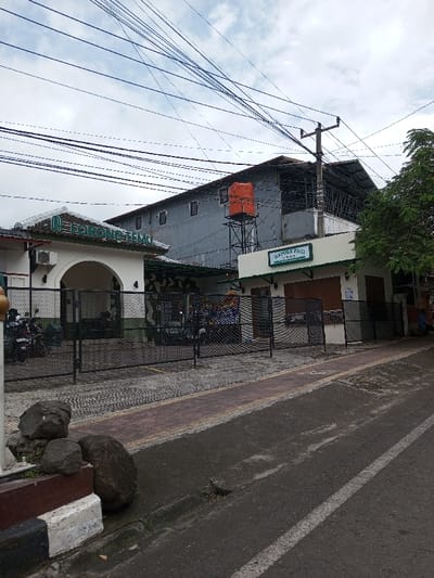 This image depicts a street scene in Vaatharawattai, Sri Lanka, under an overcast sky. The foreground features a dark asphalt road with a faded white line on the right, bordered by a concrete curb with black and white painted stripes. Alongside the curb, a paved sidewalk comprises light gray pavers with a central reddish-brown strip. Several large, dark gray rocks are positioned on the sidewalk near the left edge of the frame. A small, dark bird is visible on the street's edge near the right-hand curb.
Two primary buildings are visible behind a black mesh fence. The building on the left features a white facade with an arched entrance and green decorative trim along its roofline. A sign above the arch reads "RORONO TEMU" (partially obscured). An air conditioning unit is mounted on its exterior. Behind the mesh fence, two motorcycles are parked. The building to its right, slightly set back, is also white with a green sign that reads "DAPOER PAGI." This building has a closed roll-down shutter visible. Posters with colorful graphics are affixed to the wall between these two front structures, behind the fence.
In the background, a large, multi-story grey building with multiple rectangular windows is visible, possibly undergoing construction or renovation, with exposed metal framework on its upper right section. An orange cylindrical water tank, supported by a black metal tower, stands prominently in front of this large grey building. A utility pole with multiple cross-arms and numerous tangled overhead electrical wires dominates the upper central and right portions of the frame. A green leafy tree is partially visible on the far right. No individuals are present or actively engaged in the scene.