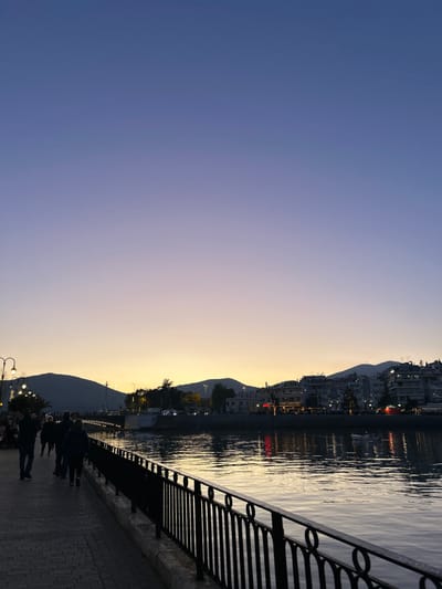 The image depicts a vibrant waterfront scene in Chalkida, Greece, at dusk or early evening. The sky, filling the upper portion of the frame, displays a striking gradient from deep indigo-blue on the left to a soft lavender and warm, pale yellow-orange on the right, indicative of a sunset or sunrise. Below this colorful sky, a tranquil body of water reflects the fading light and distant city.
Along the far bank, a series of buildings, appearing as a town or city, are built into the gentle slopes of the mountains that rise behind them. Many of these structures show illuminated windows and balconies, casting warm glows that shimmer on the water's surface. The mountains themselves are dark silhouettes against the luminous sky.
In the foreground, a paved promenade runs parallel to the water's edge. A distinctive black metal railing, featuring repeating circular designs, lines the walk. Several streetlights are visible along the promenade, some casting illuminated circles on the pavement. Scattered across the lower right of the promenade are dark silhouettes of several people, either in groups or alone, leisurely strolling. The scene evokes a peaceful and relaxed atmosphere characteristic of an evening walk by the water. No text is visible in the image.