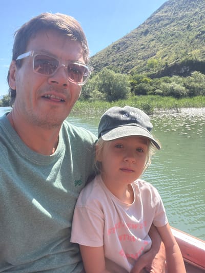 An adult male and a young girl are depicted in a selfie-style photograph, seated in a reddish-brown boat on a body of water in Vranjina, Montenegro. The man, positioned behind the girl and slightly to her left, has fair skin, dark hair, and stubble, and wears clear-framed sunglasses that reflect the person holding the camera. He is wearing a light green crew-neck sweatshirt with a small green "Superdry" logo on the left chest. His mouth is slightly open, revealing his upper teeth and tongue. His right arm is around the girl. The girl, facing the camera, has fair skin and light hair, and wears a dark grey/blue baseball cap with "LOS ANGELES VINTAGE ROOTS" written in light text. She is wearing a light pink t-shirt with partially visible pink text, "STR[AWBERR]Y WITH [CREAM]". She looks directly at the camera with a neutral expression. The water, a light green/teal color, shows ripples and sunlight reflections, and extends into the background where reeds and other lush green vegetation line its banks. Behind the vegetation and water, a large, steep, green mountain covered in trees and foliage rises under a clear blue sky, visible in the upper background, particularly on the right side of the frame. The scene is brightly lit by natural sunlight.