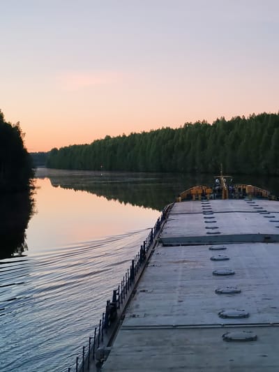This media file presents a serene landscape featuring a large, flat-decked vessel, likely a barge, navigating a calm river or canal. The photograph is taken from the vessel's deck, looking forward and slightly to the port side.

The deck of the vessel, light gray in color and adorned with rows of circular hatches and a dark railing, occupies the foreground and right side of the image. Towards the bow, a small superstructure with yellow elements and various pieces of equipment is visible.

To the left, the tranquil water beautifully reflects the sky's soft, pastel hues—pinks, oranges, and pale blues—as well as the dense, dark green trees that line both banks of the waterway. A gentle wake trails from the moving vessel on the left side, indicating its steady progress. The thick forest creates a natural, verdant corridor through which the river flows.

The clear sky and still water, with their warm, diffused colors, strongly suggest either sunrise or sunset, imbuing the scene with a pervasive sense of peace and quiet. No specific text is visible in the image. The setting for this peaceful journey is Devyatiny, Russia.