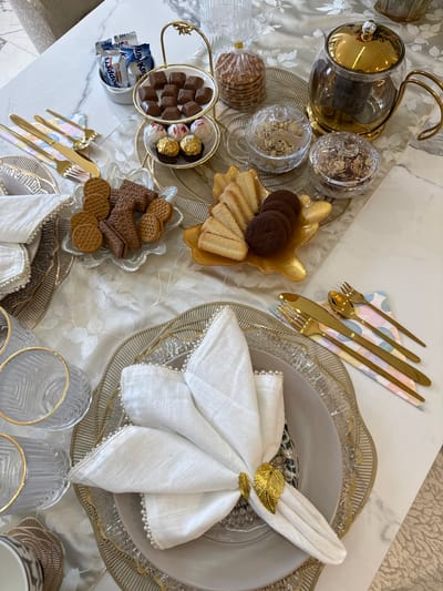 A high-angle shot shows a table setting with snacks and tableware. A clear teapot with a gold lid and handle sits to the left of two clear jars filled with food. Next to these is a stack of packaged cookies. A two-tiered serving dish holds chocolates, chocolate candies, and mini candy bars. A gold leaf-shaped bowl is filled with cookies. Two sets of gold-colored cutlery are arranged on patterned napkins. A dinner plate with a white cloth napkin and a gold leaf napkin ring is placed in the center of the image. Two gold-rimmed drinking glasses are placed at the bottom center. A runner is placed between the objects on the marble table. This table is located in an unknown location.