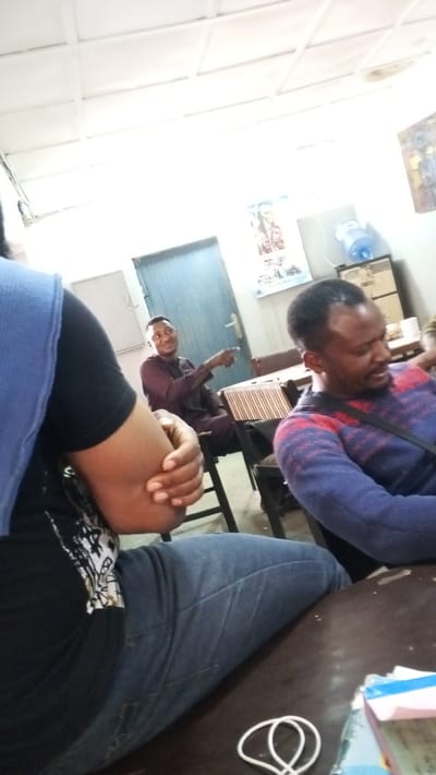 The image depicts an indoor gathering in the city of Dakata, Nigeria. Three individuals with dark skin are visible, seated at dark wooden tables. In the immediate foreground, partially obscured, is a person wearing a black t-shirt under a blue sleeveless top and blue jeans, with their right arm bent. In the mid-ground, a man in a dark maroon long-sleeved tunic is seated, smiling and pointing his right index finger to the right. To his right, another man in a blue, red, and white striped long-sleeved sweater is seated, looking downwards and to his right.
The environment features light-colored walls and a white grid-patterned ceiling with fluorescent lighting. A light blue door with a metallic handle is visible on the left side of the background. A poster featuring human figures is affixed to the wall behind the man in maroon. On the right, a blue water dispenser bottle rests on a cabinet or shelf, next to a partially visible framed item on the wall. A white coiled cable and a stack of papers with a blue cover are present on the foreground table. The seating arrangement suggests a meeting, classroom, or communal space.