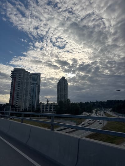The image depicts a cityscape with modern high-rise buildings set against a dramatic sky filled with textured clouds. In the foreground, a concrete barrier and railing suggest the perspective is from an elevated roadway or bridge. Several lanes of traffic are visible below, with cars moving along the highway. The buildings are contemporary in design, featuring glass facades and balconies. Trees and other greenery are interspersed between the buildings and along the roadside, indicating a suburban or urban fringe environment. The sky is a mix of bright white and dark gray clouds, with the sun partially obscured, creating a diffused light. The overall impression is that of a vast, urban landscape under a dynamic, cloud-covered sky. Based on the architecture and the presence of coniferous trees, this scene is likely in the District of North Vancouver, Canada, specifically along a major arterial route like Highway 1, with buildings in the Ironwood or Capilano areas.