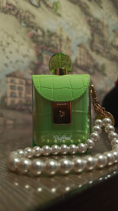 A green, purse-shaped perfume bottle with a textured flap and a gold clasp sits on a wooden surface. A string of pearls is draped around the bottle and extends into the foreground, casting reflections on the surface. The bottle has the brand name "Puffmi" printed on its lower front. The background is a blurred image of buildings and foliage, suggesting an outdoor or decorative setting, possibly a tapestry or mural. The scene appears to be an indoor still life. There is no indication of Zvenigorod, Russia.