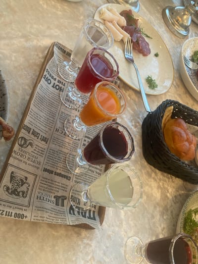 The image shows a close-up view of a table setting, likely at a meal or gathering. The scene is set indoors, possibly in a restaurant or dining room, with a textured, light-colored tablecloth.
On the table, several objects are visible. In the foreground, there are four small glasses containing colorful liquids, appearing to be juices or aperitifs. These glasses are arranged on a wooden tray lined with what looks like newspaper clippings. To the left, a plate holds slices of cured meat and some white food items, possibly cheese or fatback, with a fork resting on it. In the background, a woven basket contains a small, golden-brown bread roll. Another plate with some greenery is also partially visible. A glass of dark red liquid, likely wine, sits to the right.
The focus is on the drinks and food, suggesting a tasting or a starter to a meal. The newspaper clippings under the glasses add an interesting, possibly decorative, element. Some text is visible on the clippings, including "HOT OFF THE PRESS," "SUPER GROW FERTILISERS," and "+000 111 22." The overall impression is one of a well-prepared meal or event, with attention to detail in presentation. There are no people visible in the frame. The lighting suggests it might be daytime, possibly indoors with artificial lighting.