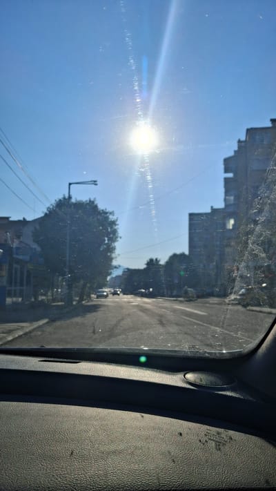The image is a view from inside a vehicle, looking through the windshield. The sun is high in the sky, causing a strong lens flare that obscures parts of the scene. The windshield is dirty, with visible streaks and dust.
The scene outside the car is a city street with buildings and trees. On the left, there are storefronts. In the center, a tree stands next to a streetlight, and cars are visible on the road. On the right, a multi-story building dominates the skyline. The road appears to be asphalt. A green light is visible on the right side of the road.
The interior of the vehicle shows the dashboard, which is dark grey and textured. A small, round speaker is visible on the dashboard.
The location is identified as the city of Gotse Delchev, Bulgaria, on a clear, sunny day.