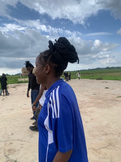 The image depicts a group of people outdoors in Gwafan, Nigeria. The scene appears to be a wide, open, paved area with a grassy field and some distant buildings in the background. The sky is partly cloudy with dramatic, billowing clouds against a bright blue sky, suggesting it might be daytime, possibly late morning or afternoon.
In the foreground, a young woman is captured from the side, her head bowed and turned to her left. She has dark hair styled in braids, tied up with a black ribbon. She is wearing a vibrant blue t-shirt with white stripes on the sleeves. Her expression is not clearly visible, but her posture suggests focus or contemplation.
Several other individuals are visible in the mid-ground and background, some appearing to be engaged in activities or standing around. One person in the background is wearing a white garment. Another group of people further back includes individuals in dark clothing, and one person in a white shirt and blue jeans. The ground beneath them is a light-colored, textured surface, possibly concrete or stone.
The overall atmosphere seems relaxed and casual, with people gathered in an outdoor space. The weather appears to be pleasant, with the dynamic clouds adding visual interest to the sky. There is no visible text in the image.