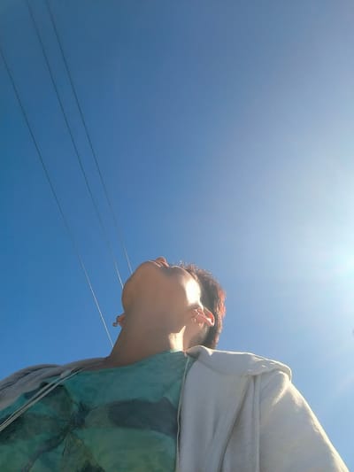 A low-angle, close-up shot captures a person looking upwards into a clear, bright blue sky on a sunny day. The sun is prominently visible on the right side of the frame, casting a bright glare. Three dark, parallel lines, likely power lines, extend diagonally from the upper left corner of the image into the sky.
The person, whose face is partially obscured as they tilt their head back, appears to be wearing a teal-colored t-shirt with a dark graphic print, possibly a palm tree. They are also wearing a light-colored hooded jacket over the t-shirt. Visible jewelry includes earrings on both ears and a ring on one finger. The person's hair is a reddish-brown hue.
The overall impression is one of outdoor activity, possibly in a park or open area, given the clear sky and power lines. The bright sunlight suggests it is daytime, likely late morning or afternoon. The mood conveyed is one of peacefulness and enjoyment of the outdoors. No text is visible in the image.