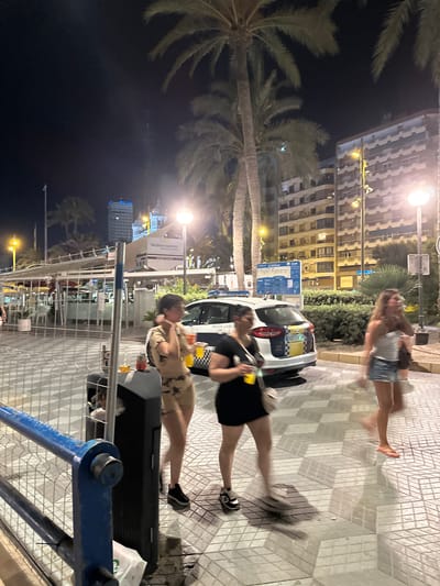 This nighttime, full-frame, eye-level shot depicts a street scene in Alicante, Spain, likely along the Explanada de España, characterized by palm trees and illuminated buildings. In the foreground, a blue metal railing runs horizontally across the bottom of the frame. To the right of the railing, a black trash receptacle stands partially obscured.

Three women are the primary human subjects. The woman on the far left, wearing a patterned two-piece outfit and black sneakers, is captured mid-motion, holding a yellow drink. Next to her, a woman in a black dress and sneakers is also in motion, carrying a purse and a yellow drink. On the right, a third woman in a light-colored top and denim shorts is walking away from the camera, her movement blurred.

A white police vehicle with blue and yellow markings, identified as "POLICIA LOCAL," is parked on the right side of the image, its rear facing the viewer. Behind the women and the police car, a patterned tiled sidewalk is visible. Beyond the sidewalk, a street lamp illuminates the scene, and several buildings, including a tall, modern structure with "GVA ELS" text, are visible in the background, along with more palm trees. The overall atmosphere is that of a lively urban environment at night.