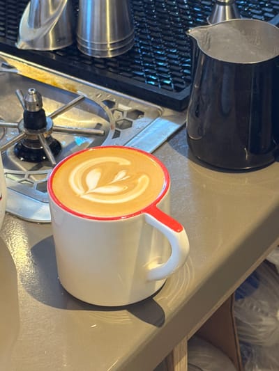 A close-up, eye-level shot shows a white ceramic mug with a red rim and handle, filled with latte art, on a grey countertop. The latte art appears to be a rosetta pattern. To the left of the mug, a chrome espresso machine part with spokes is visible. Behind the mug, a black stainless steel milk pitcher is positioned on the countertop. In the background, out of focus, are stainless steel containers and a dark, textured surface, possibly a wall or backsplash. The lighting suggests an indoor environment, likely a cafe or bar. The image is taken in West Hollywood, California, United States of America.