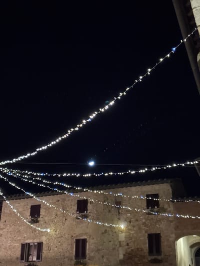 Night scene showing illuminated string lights hung diagonally against a dark sky. To the right, a building with a stone facade and dark-shuttered windows is visible. Additional strands of lights are draped vertically along the building's facade. A light source, possibly the moon, shines faintly between the strands, illuminating the tower. The top portion of the image shows sections of two buildings.
