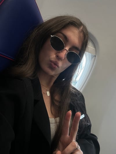 A young woman with long, dark brown hair is captured in a close-up selfie, reclining in what appears to be an airplane or vehicle seat. She wears dark, round sunglasses with a silver frame and has her lips slightly pursed in a pout. Her left hand is raised, making a peace sign, and a silver ring with a small stone is visible on her ring finger.

Around her neck, she wears a silver chain necklace featuring a rectangular, dog-tag-style pendant with faint, illegible text, alongside a small, gold-colored charm. She is dressed in a black blazer or jacket worn over a light-colored top, possibly ribbed.

The background shows an interior space, likely a cabin, with a window revealing bright daylight. The seat she occupies is dark blue with a reddish-pink trim. On a piece of black material, possibly part of her jacket or a bag, a reflective label clearly displays the text "MADE IN ITALY". The overall scene suggests a relaxed moment during travel. The photo is associated with Dortmund, Germany, implying travel to or from this location.