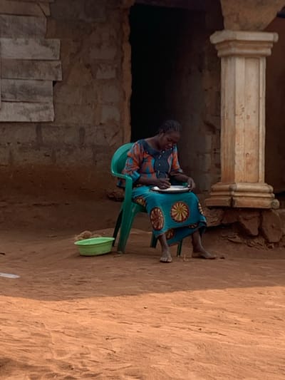 An adult female is seated outdoors on a green plastic chair on reddish-brown, dry earth. She has dark braided hair and is wearing a long-sleeved patterned top with orange, blue, black, and white elements, and a long blue skirt with yellow, red, and black circular patterns. She is barefoot. She holds a white plate in her lap and is looking down, engaging with its contents with both hands; a dark object is visible in her right hand, suggesting eating or preparation. To her left, on the ground, is a small light green plastic basin. Behind her, a rustic structure with light brown block walls is visible, featuring a dark, open doorway in the center and horizontally placed weathered wooden planks on the far left. To the right, a light-colored concrete column with a tiered base and cap stands. This scene is located in Aguleri, Nigeria.