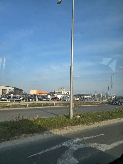 This image captures a daytime scene along a roadside in Stroevo, Bulgaria. In the foreground, a section of asphalt road exhibits faded white directional markings, possibly indicating a turn. To the right, an unkempt strip of green and brown grass runs parallel to the road. A prominent, tall, slender grey metallic lamppost with a modern fixture at its apex is centrally positioned on this grassy strip.

Beyond the grassy verge, a multi-lane asphalt road or highway is visible, separated by a metal guardrail. A dark-colored sedan, possibly a BMW, is observed on the far right lane of this road, traveling away from the viewer.

The background reveals an expansive commercial or industrial complex. Numerous vehicles, predominantly utility vans and trucks, are parked in orderly rows across multiple open-air lots. Several commercial buildings are present, including a two-story structure with a light-colored facade to the far left, and a building with a distinctive orange roof further right. Additional streetlights and utility poles are distributed throughout the background area.

The sky overhead is predominantly clear and blue, with sparse, wispy white clouds. Reflections of an interior space are visible on the upper left and right sections of the image, superimposed over the sky, suggesting the photograph was taken through a window.