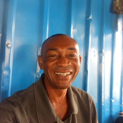 A single adult male, appearing to be of African descent, with dark skin and a very short, dark haircut, is depicted in a frontal, head-and-shoulders portrait. He is smiling widely, revealing his upper and lower teeth which exhibit some discoloration. He wears a grey, textured, collared shirt. The background is composed of a vibrant blue corrugated metal surface, featuring distinct vertical ridges and visible circular metallic fasteners or rivets, particularly on the left side of the frame. The lighting is bright, creating a sheen on the man's forehead. This scene is situated in the city of Koko, Nigeria.