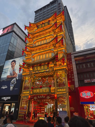 The image shows a street scene in Jinjiang District, China. A multi-tiered, brightly lit, gold-colored building with Chinese characters and red lanterns is prominent. A modern building with a large Longines advertisement featuring a man and a watch is visible below. People are walking on the street in front of the buildings. Multiple Chinese signs in red and gold are present.