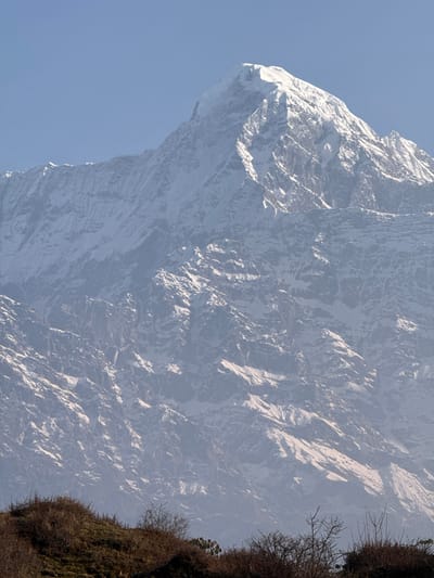 A striking view of a snow-covered mountain is captured in Lwang Ghalel, Nepal. The mountain dominates the frame, its rugged, steep slopes covered in patches of snow and rock. The peak extends to the left side of the frame, silhouetted against a light blue sky. In the lower right corner, a small area of vegetation clings to a rocky cliff, adding a touch of green and brown to the scene. The overall impression is one of a remote, mountainous landscape.