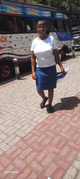 This outdoor photograph, taken in what appears to be Biashara ward, Kenya, shows a person standing next to a bus on a sunny day. The central subject is an adult, possibly female, with dark skin and short dark hair, wearing a white short-sleeved top, a dark blue skirt, and dark flat shoes. She is holding a blue object or bag in her hands and has a slight smile while looking towards the camera.

To her left (viewer's left), a large passenger bus is parked, occupying a significant portion of the background. The bus is predominantly blue and white, adorned with colorful, rainbow-like stripes and various designs. Partially visible text on the bus includes "Infinix" near the top and other less legible stylized text and patterns on its side. The front wheel of the bus is visible in the lower left.

The ground is paved with red and grey interlocking bricks laid in a herringbone pattern. Strong, elongated shadows cast by the person and other unseen objects indicate bright sunlight, suggesting it is daytime. The overall setting appears to be a street, bus stop, or a parking area for public transport. A watermark that reads "police kombo" with a small pink graphic is visible in the bottom right corner of the image.
