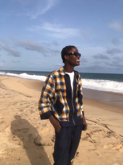The image depicts a man on a beach. The man is Black, wearing dark sunglasses, a white shirt, a plaid jacket with a zipper, and dark pants. He is looking upward and smiling. The background features a beach with sand, ocean waves, and a sky with some clouds.
Based on the prompt, the location is Epe, Nigeria, though this is not discernible from the image.
