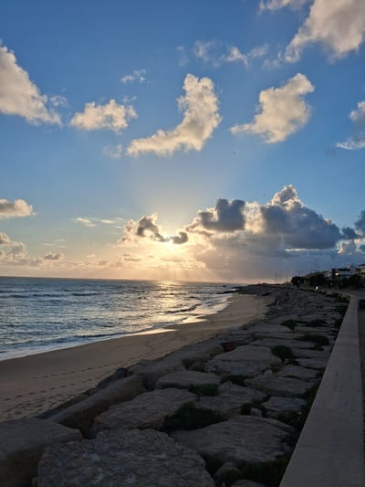 The image shows a beach scene in Figueira da Foz, Portugal. The view is rotated, presenting the sky on the left and the sea on the right. Above, a blue sky is dotted with white and gray clouds, some obscuring the sun. Birds are visible flying. The sea reflects sunlight, creating a shimmering effect. A beach runs between the sea and a stone embankment. Above the embankment, there is a sidewalk and buildings in the distance.