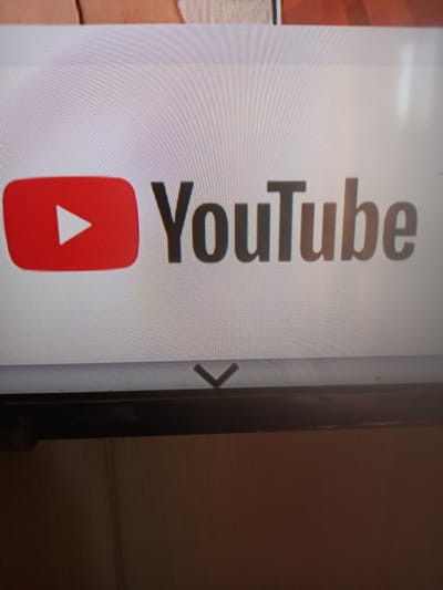 The image displays a close-up view of the YouTube logo, which consists of a red play button icon next to the word "YouTube" in dark grey lettering. The logo appears on a screen, with the pixelated texture of the display visible. Below the logo, a downward-pointing chevron icon is present, suggesting a navigation or expansion element. The screen itself is framed by a dark border at the bottom. The background above the screen appears to be a blurred image of a wooden surface. The image is taken at an angle, and the lighting suggests an indoor environment, possibly a room. There are no people or animals present in the image. The overall scene is static and focuses solely on the digital display of the YouTube logo. There is no information within the image to suggest its location in Adutiškis, Lithuania.