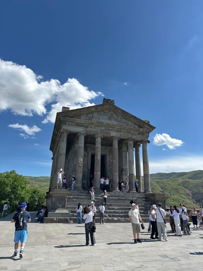 In Garni, Armenia, the Temple of Garni stands against a backdrop of rolling, verdant hills under a bright blue sky dotted with fluffy white clouds. The ancient Greco-Roman temple, constructed from gray stone, features a prominent facade with a triangular pediment and a series of Doric columns. A wide, stone staircase leads up to the temple's entrance, where several people are gathered.

The foreground is a large, paved plaza. A young man in a blue baseball cap, navy t-shirt, and light blue shorts with a backpack walks away from the camera towards the temple. To his right, a woman in a white t-shirt and dark pants stands looking towards the temple.

Numerous other individuals, mostly dressed in casual, warm-weather attire, populate the scene. Some are ascending the temple stairs, while others are standing on the plaza, observing the structure or interacting with each other. The overall atmosphere is one of tourism and exploration, with visitors admiring the historical site. The natural landscape surrounding the temple adds to the picturesque quality of the scene.