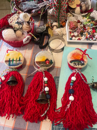 The image shows a close-up, overhead view of a craft project featuring three handmade ornaments. Each ornament has a circular, felted top depicting a landscape scene with a sky, land, and details like trees, a house, and a fence. Below the felted top, each ornament has a thick tassel made of red yarn, resembling hair. Various beads in white, blue, green, and red are strung along the top of the yarn tassels, and two of the ornaments have small, brass bells attached. The ornaments are laid out on a checkered tablecloth. In the background, various craft supplies are visible, including spools of yarn, scissors, lace, beads, and small decorative items, suggesting a crafting area. The image is described as being taken in Cheshnegirovo, Bulgaria, however, no specific city features or landmarks are identifiable within the photograph to confirm this location.