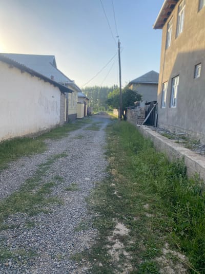 The image depicts a rural dirt road in Боғистон, Tajikistan, lined with residential buildings. On the left, a long, white-walled building with a corrugated metal roof is visible. To the right, a multi-story concrete building with white-framed windows dominates the foreground. Further down the road, a smaller house with a grey, pitched roof and yellow siding can be seen. The road surface is composed of gravel and dirt, with patches of overgrown grass along the sides. A wooden utility pole with wires stands on the right side of the road. In the background, trees and more residential structures suggest a village setting. The sky is clear and blue, indicating daytime. There is no visible activity or interaction depicted in the image.
