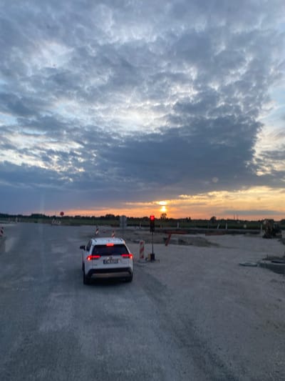 The image captures a sunset over a construction site in Podłęże, Poland. A white SUV is stopped at a red traffic light, facing away from the camera. The road surface is gravel and unfinished. Construction barriers, including striped poles and a stop sign, are visible. In the distance, trees line the horizon, and the sky is filled with dramatic, cloudy formations tinted with the warm hues of the setting sun. A construction excavator is also visible on the right side of the frame. The overall impression is one of an active, developing area at dusk.