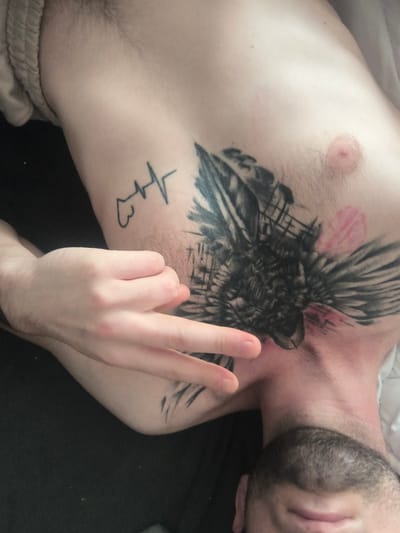 The image shows a close-up, high-angle shot of a person's torso and neck, lying down. The person has multiple tattoos on their chest and upper arm, including a large black and grey design that appears to be a bird or animal with wings and a fragmented background, and a simpler line drawing of a heartbeat with a heart shape below it. The person's left arm is raised, with their hand positioned in front of their chest, fingers extended in a peace sign gesture. There are faint pink marks on the chest, possibly a temporary transfer or faint blush. The person has dark stubble on their chin and neck. The background is dark and appears to be fabric. The image does not contain any information that would allow for its location to be determined in the city of Grójec, Poland.