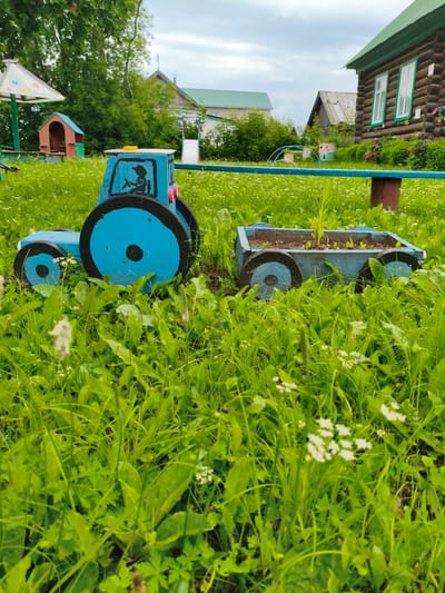 A blue wooden toy tractor with a black painted driver is in a grassy yard in the village of Munayka, Russia. The tractor has large black wheels with a black circle in the center. A smaller blue wooden wagon filled with green seedlings is attached to the tractor. In the background, there are two wooden houses with green roofs and a playground set. The sky is overcast.