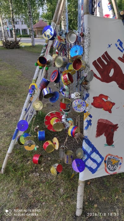 The image displays an outdoor scene in Iisalmi, Finland, likely near a park or public space, as suggested by the presence of trees, grass, and a building in the background. The main subject is a temporary, cone-shaped structure resembling a teepee, constructed from white fabric stretched over wooden poles. This structure is adorned with numerous recycled items, primarily tin cans and CDs, which are hung at various heights. The cans are colorful, some painted, and others have original labels visible. The CDs reflect the light and add a decorative element. The structure is partially supported by a wire mesh fence, to which more of the recycled decorations are attached. On the fabric surface of the structure, there are several painted elements, including what appears to be a large red handprint, a depiction of a horse's head, and a shield-like emblem with symbols and text, possibly representing a local crest or organization. The ground around the structure is covered in grass and fallen leaves. The lighting suggests it is daytime. There are no people visible in the image. The timestamp at the bottom right indicates the image was taken on July 8, 2026.