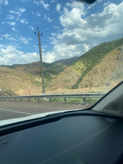 The image captures a view from inside a vehicle, looking out towards a mountainous landscape. The sky is a vibrant blue with scattered white clouds, some of which are darker and more dramatic. Power lines stretch across the sky, supported by a tall concrete utility pole. In the middle ground, a road runs alongside a metal guardrail. Beyond the road, the terrain rises into steep, arid mountains. The mountains are a mix of dry, rocky slopes and areas covered in green vegetation. A large metal pylon, likely for electricity transmission, is visible in the valley between the hills. The foreground is dominated by the interior of the car, including a portion of the dashboard and windshield. The image appears to have been taken on a sunny day, possibly in a rural or mountainous region of Chetsu, Uzbekistan, given the geological features and vegetation. There are no people visible in the scene.