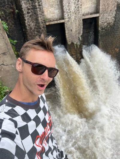 This media file captures a person taking a selfie near a powerful waterfall in Klungkung, Indonesia. The subject, a man with blond hair and a surprised expression, is wearing sunglasses and a black and white checkered t-shirt with red text. The t-shirt has a distressed look, possibly from washing. The setting is a natural landscape with a stone structure on the left, suggesting a man-made element interacting with the powerful water flow. The water is turbulent and white, indicating a strong current. The scene appears to be outdoors, possibly during the day, as there are no artificial lights visible. The man's open mouth and wide eyes convey a sense of awe or excitement in response to the powerful natural display. The stone pathway in the foreground suggests a public area where visitors can observe the waterfall. The lush greenery visible at the bottom right indicates a tropical or subtropical climate, consistent with the location in Indonesia. The stone embankment on the left seems to be part of a channel or structure guiding the water.